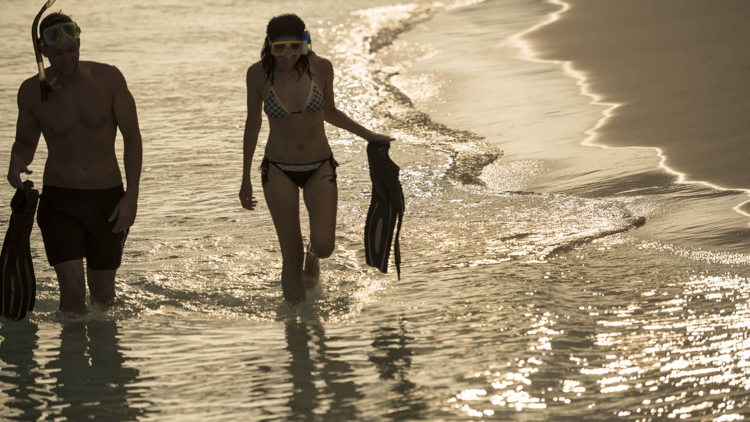 Two people in swimsuits and snorkel gear hold fins as they walk along the water's edge, sun reflecting off gentle waves.