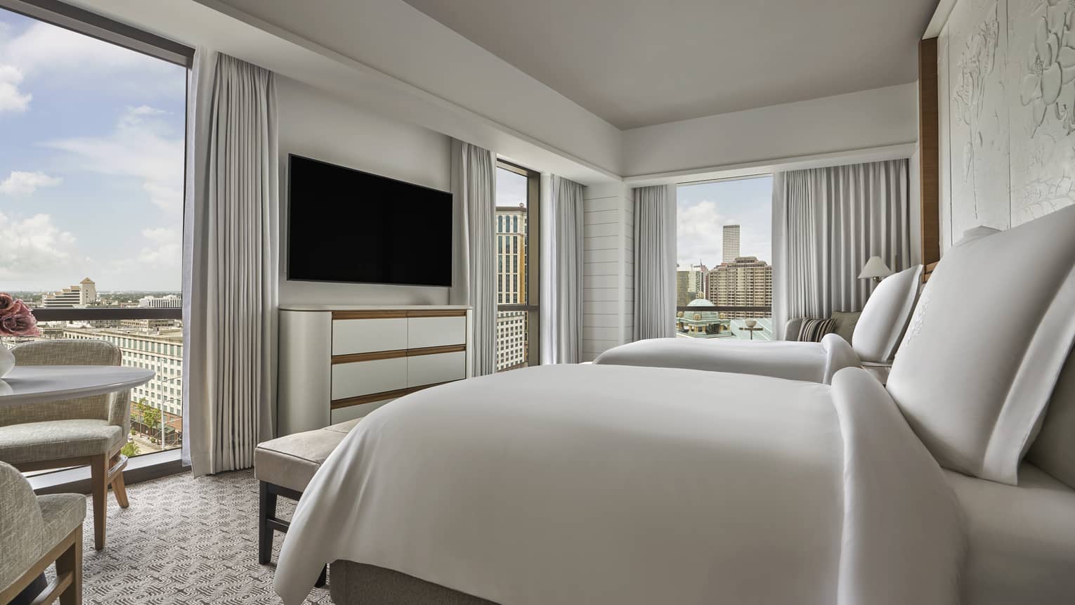 Queen bed with white linens, white curtains and chest of drawers, wall-mounted television, floor-to-ceiling windows with view of downtown New Orleans