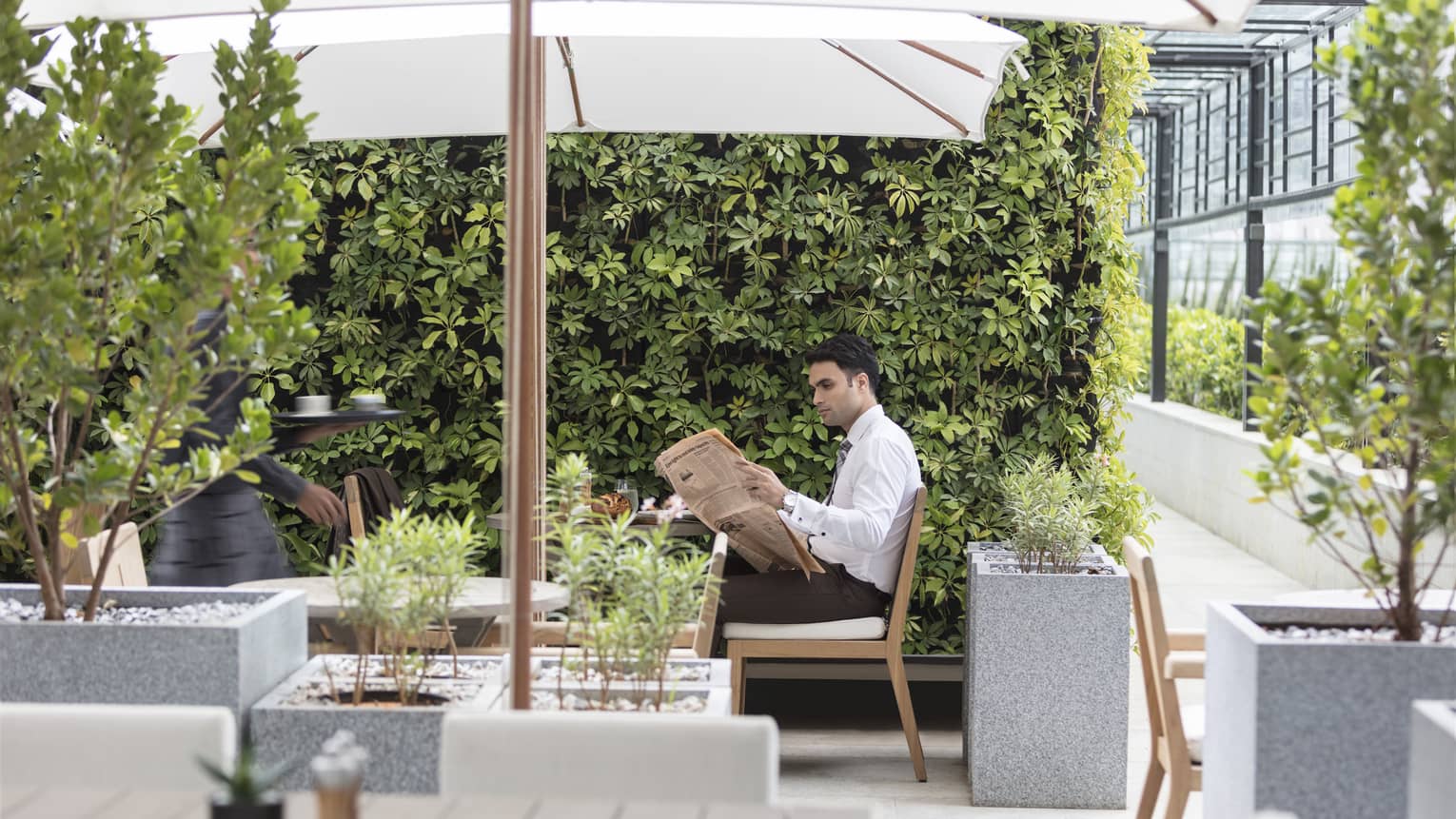 A guest reads a newspaper on an outdoor patio with foliage and umbrella covered tables.