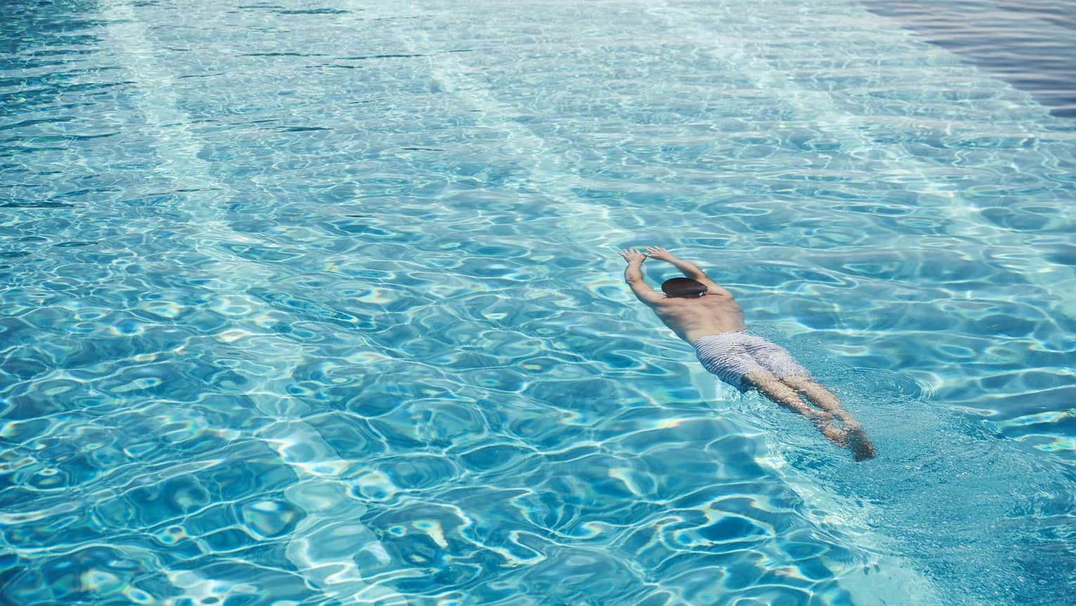 Man in swim shorts with arms outstretched dives underwater in blue swimming pool