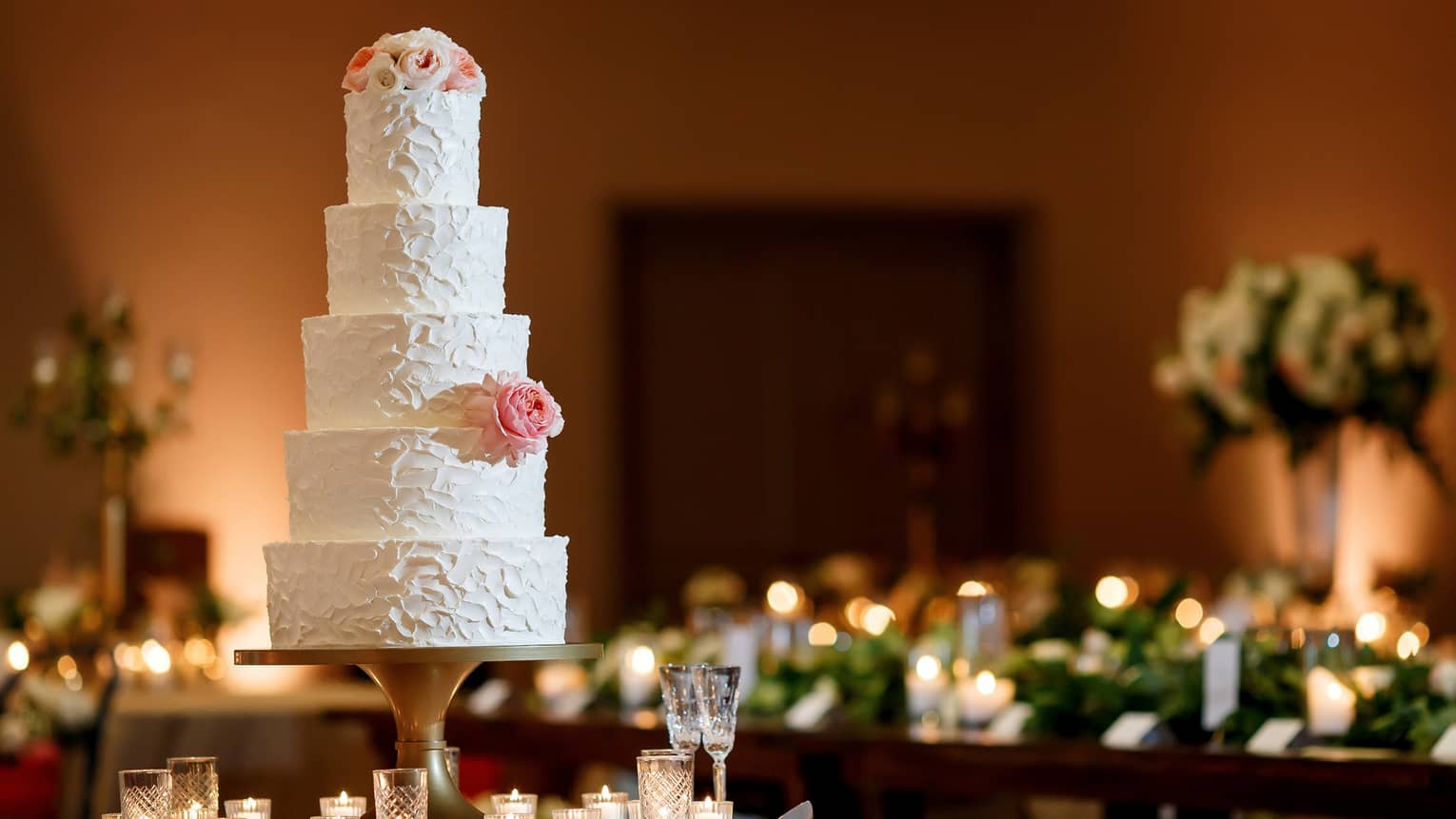 A five-tiered white wedding cake garnished with a white rose and some small white and pink flowers on the top tier , sits on a white round table surrounded by white candles