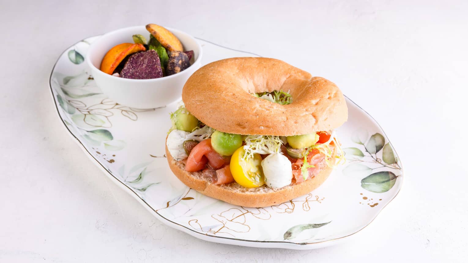 Bagel filled with sliced red and yellow cherry tomatoes, cubes of cheese and sprouts served on a floral plate with a side bowl of veggie chips