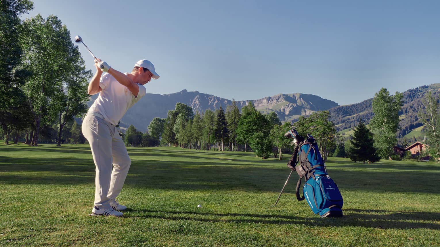 Man on mountainside golf course swings to hit golf ball beside blue golf bag