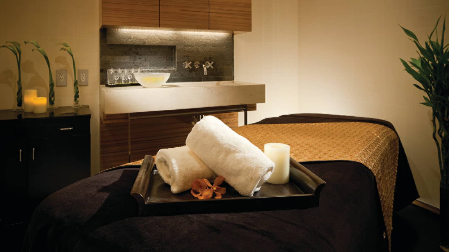 Spa treatment room, bamboo tray with towels, candles, flower on massage bed