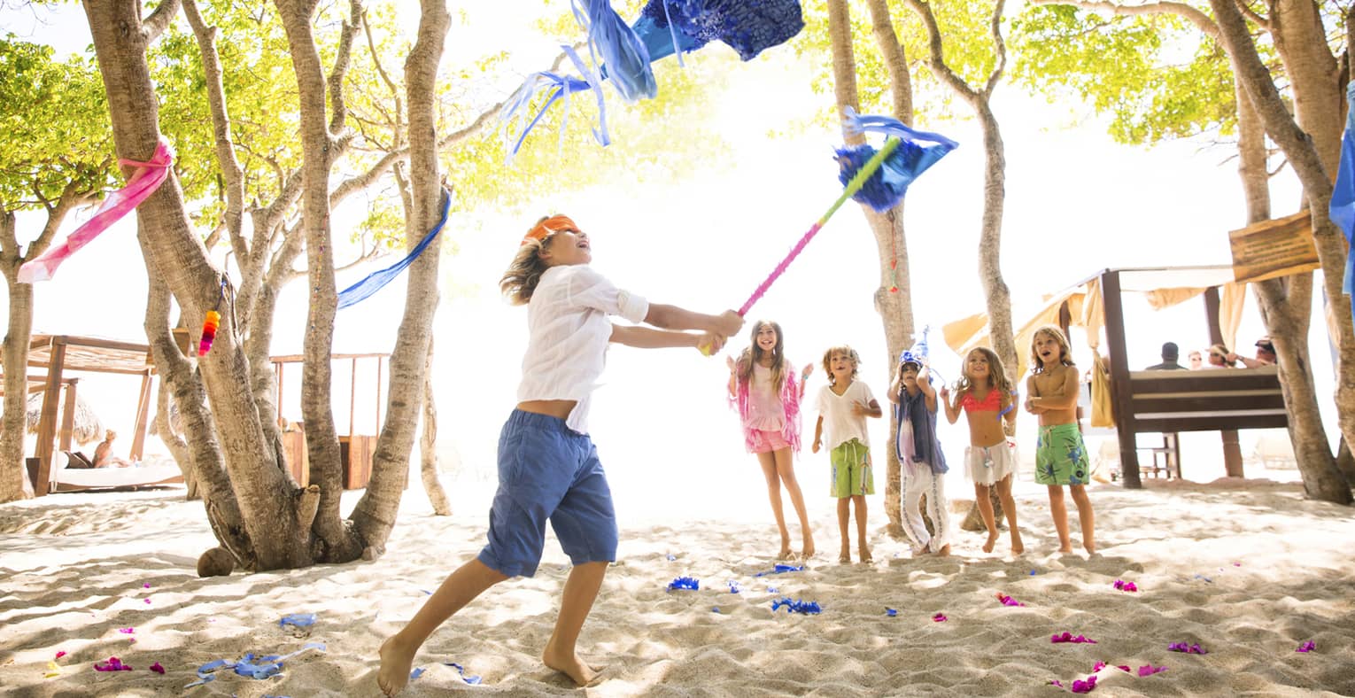 Boy wearing orange blindfold swings stick at blue pinata on beach while five young children cheer him on