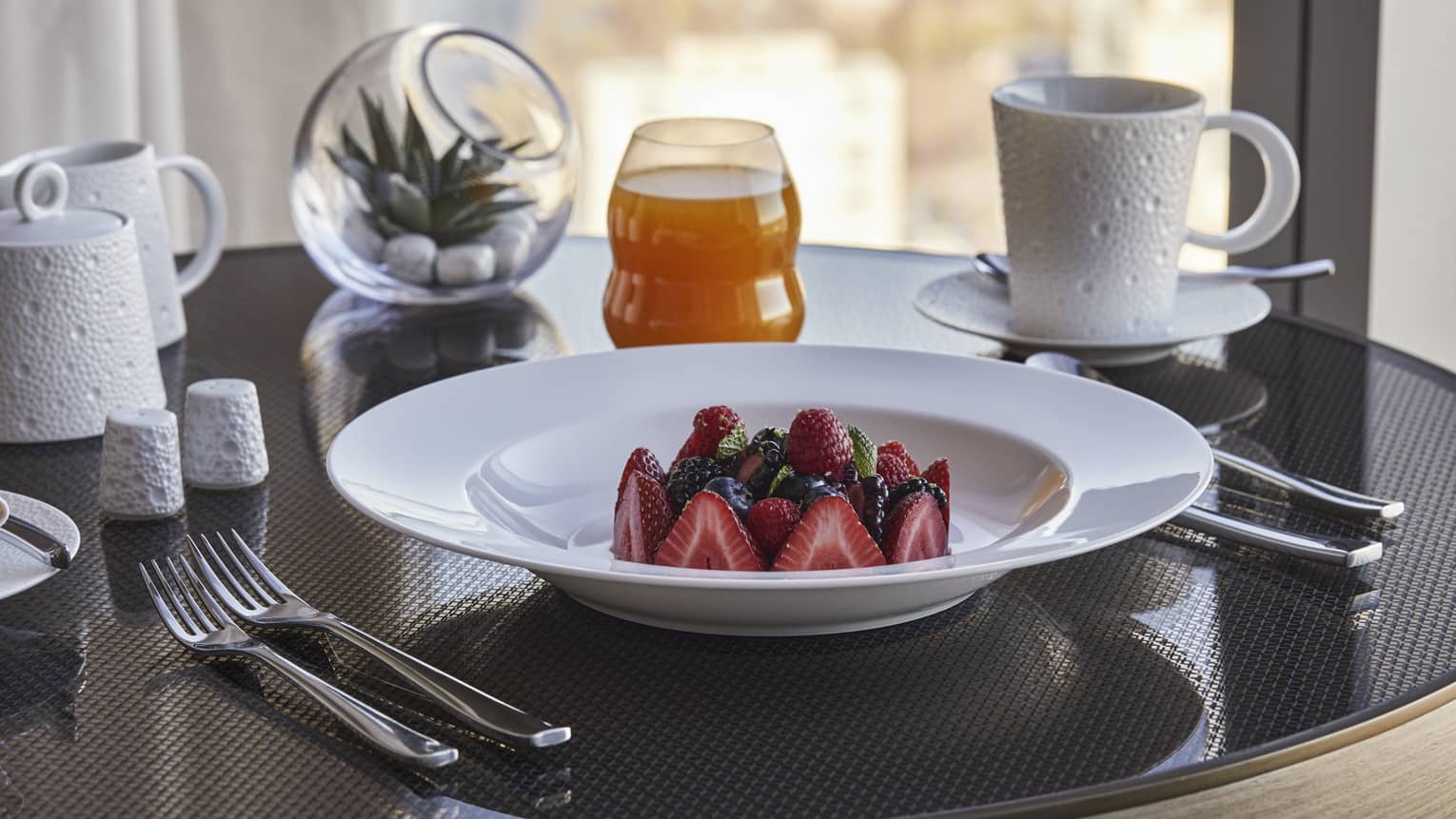 A close up in-room dining detail of strawberries and blueberries in a white porcelain bowl along with a cup of coffee and juice 
