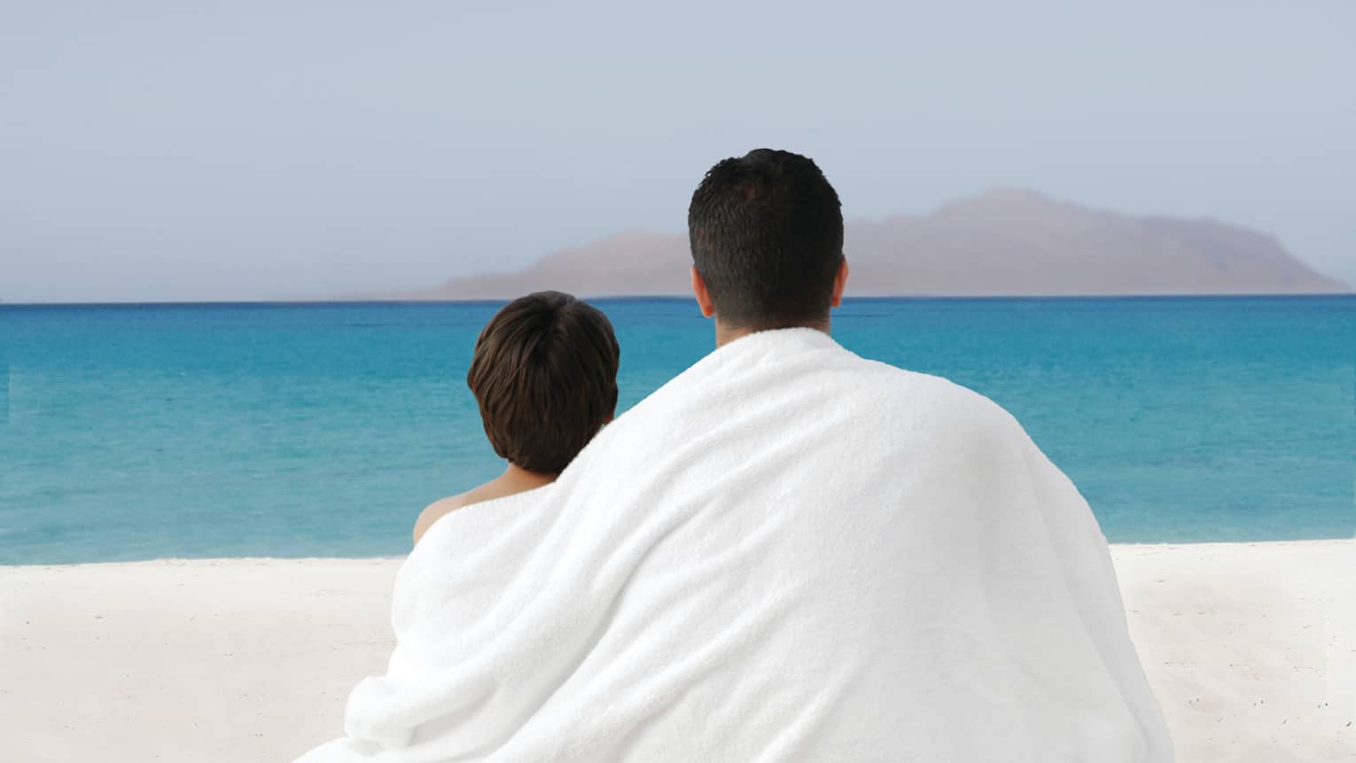 Father and son share white towel drapes across backs, look out at beach and sea