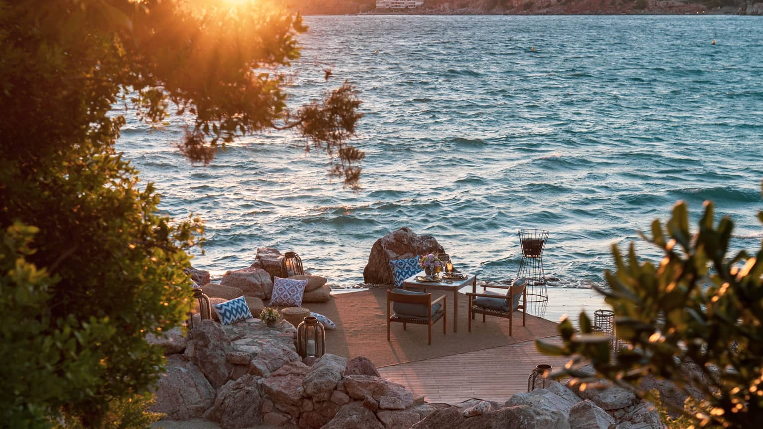 From tree-lined stairs, a furnished patio, including table set for two beside rippling water  and sun setting over a distant isle