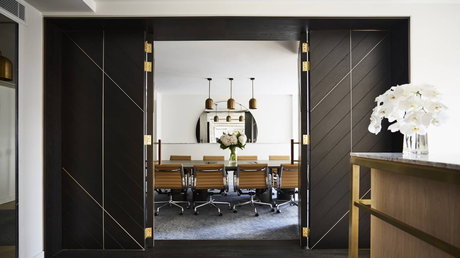 Tall open doors to private boardroom table, swivel chairs under three lights, mirror