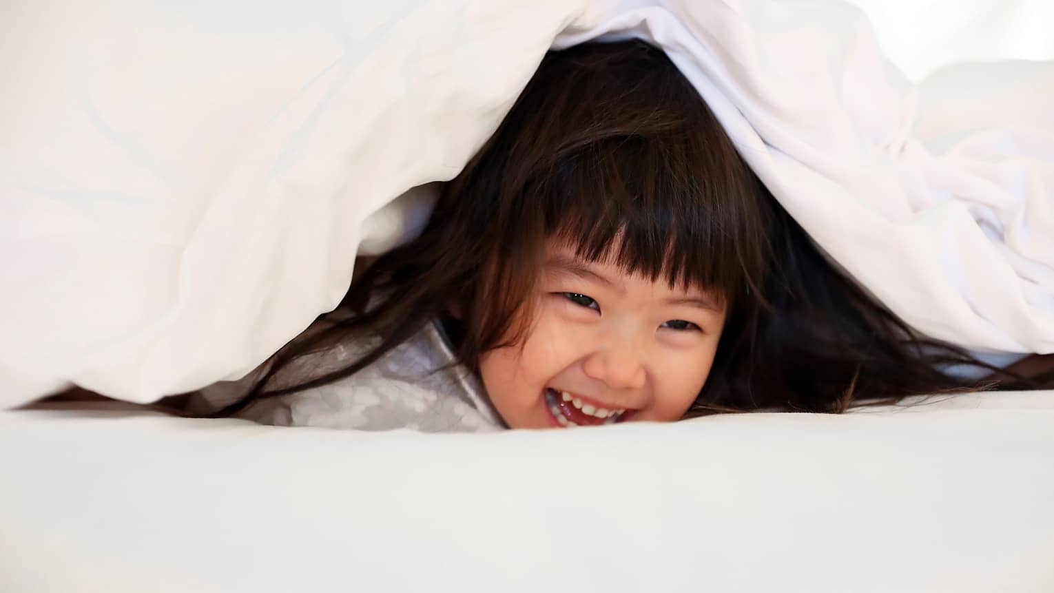 Young girl laughing as she plays under a white bed sheet
