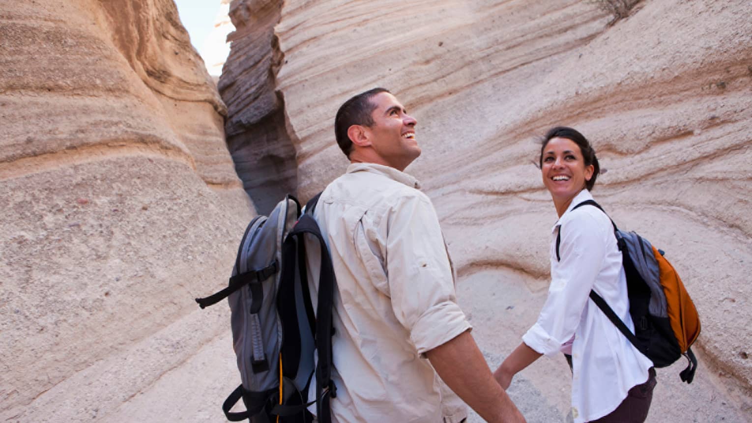 Smiling man and woman wearing backpacks hold hands, walk through desert rocks