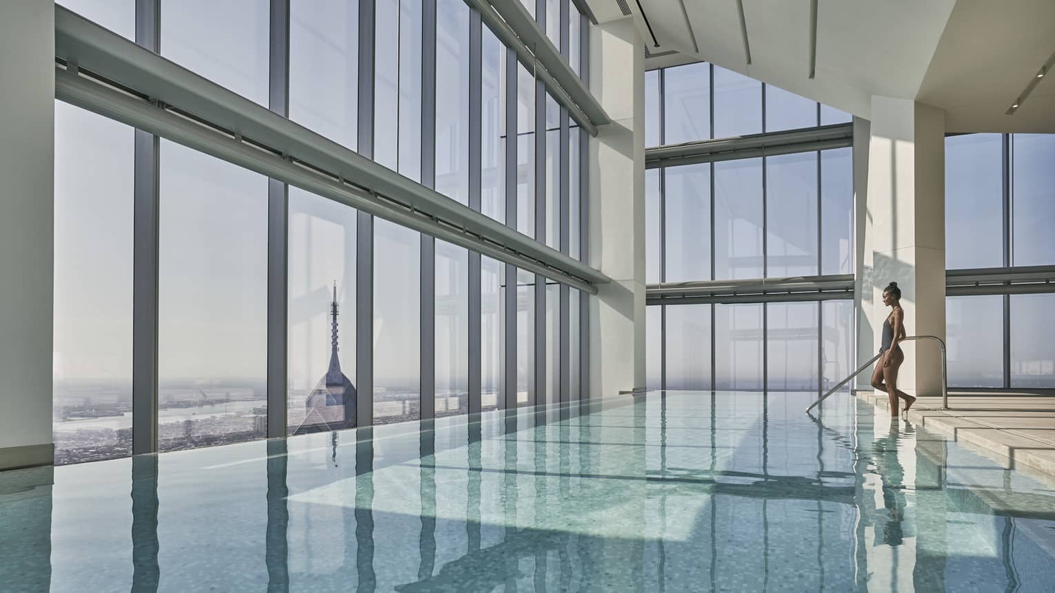 A woman enters a calm indoor pool, glass walls of windows look out to Philadelphia skyline