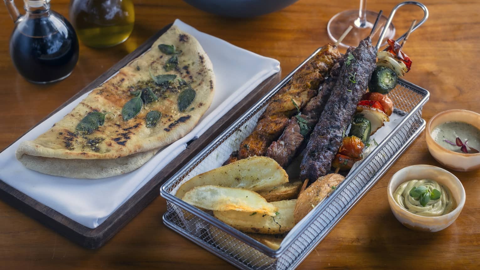 Wire basket of three grilled meat skewers served with a skewer of vegetables, pita, arugula salad, and handcut Greek potatoes