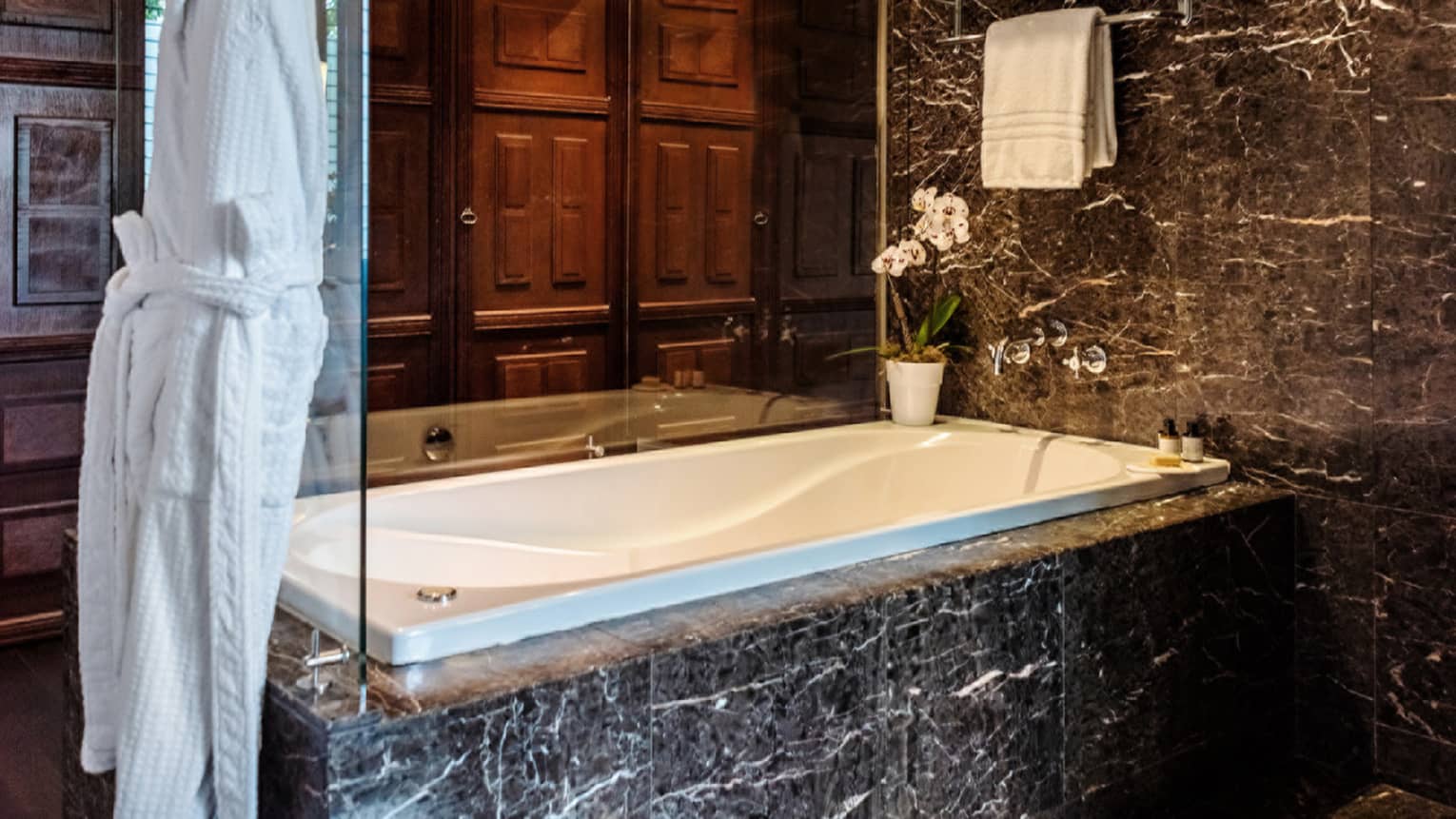 Black marble tub enclosed by glass, with hanging white bathrobe