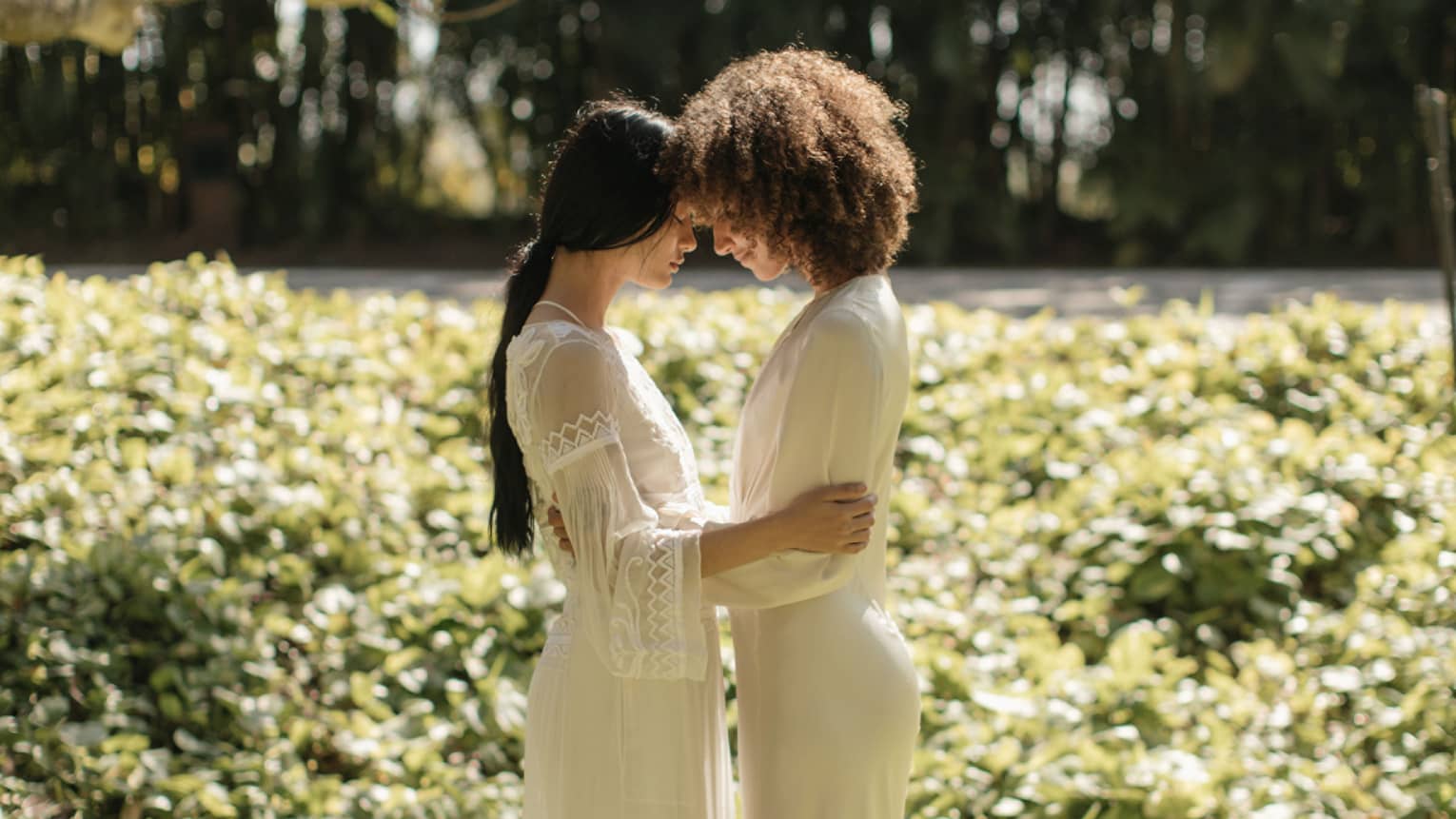 A couple of women embracing and placing their heads on each other in a beautiful field in a loving way.