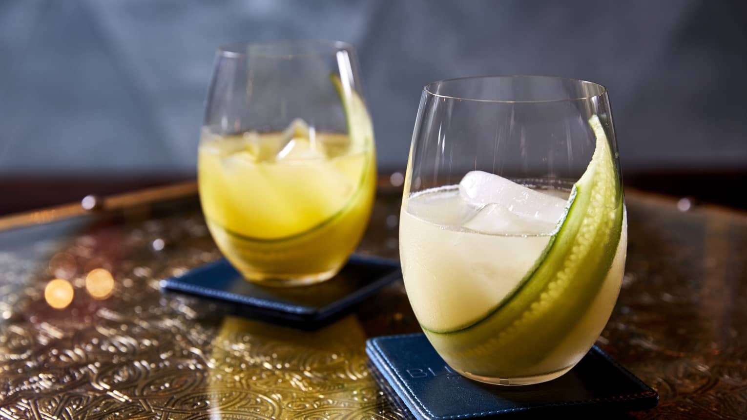 Two yellow beverages in rounded glasses with a green vegetable in the glass.