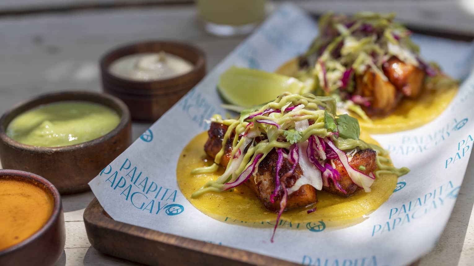 Two fish tacos sit open-faced on a paper-lined wood platter with three small wooden bowls of sauces on the side