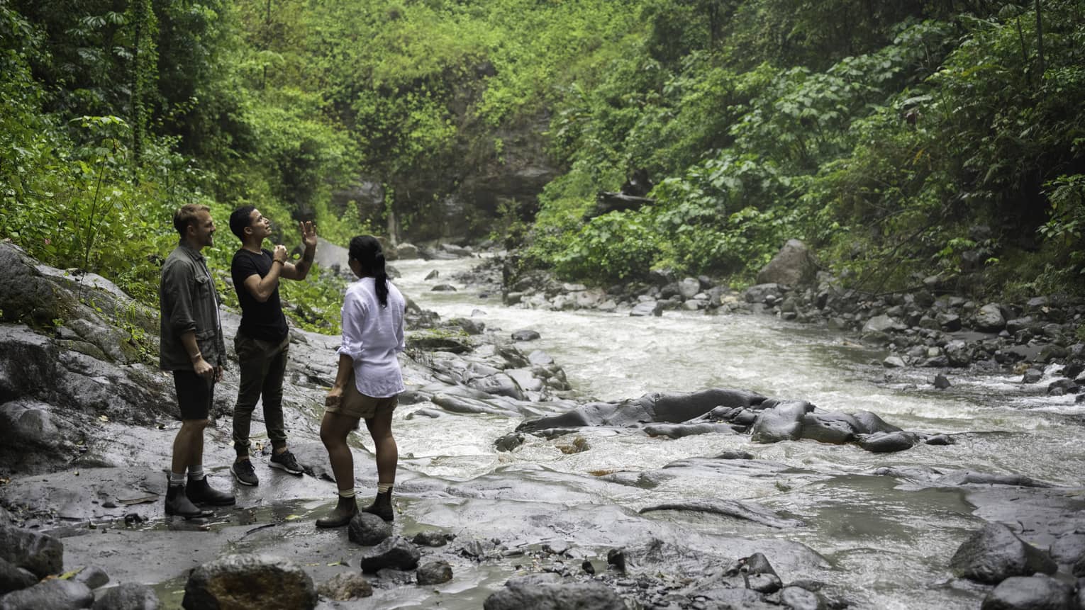 Three hikers have a chat as they stand on the bank of a rushing river surrounded by the exquisite green of lush rainforest. 