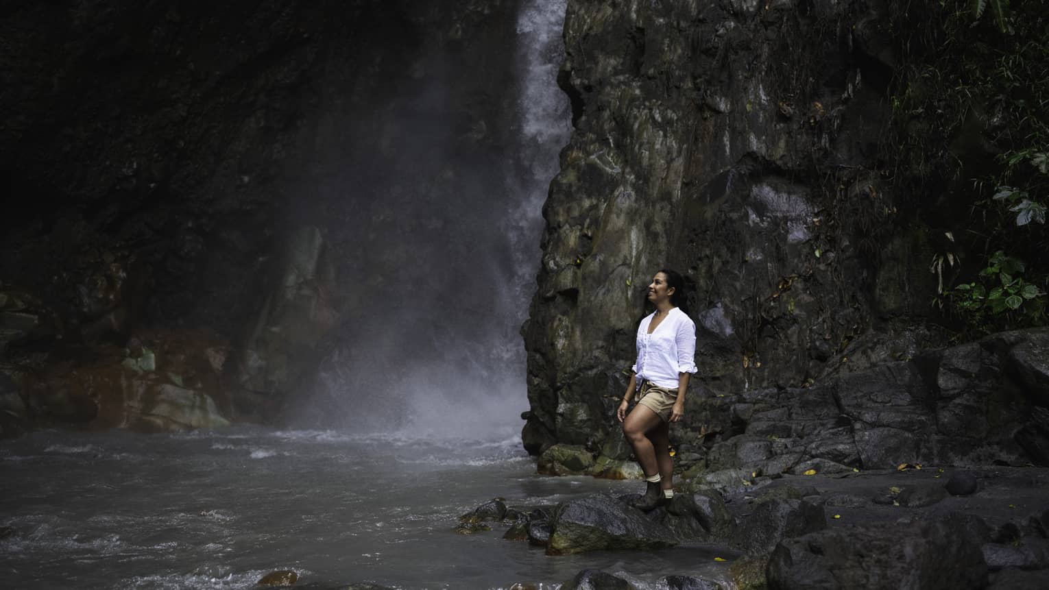 ,Woman wearing white shirt and brown shorts stands on a rock next to a waterfall