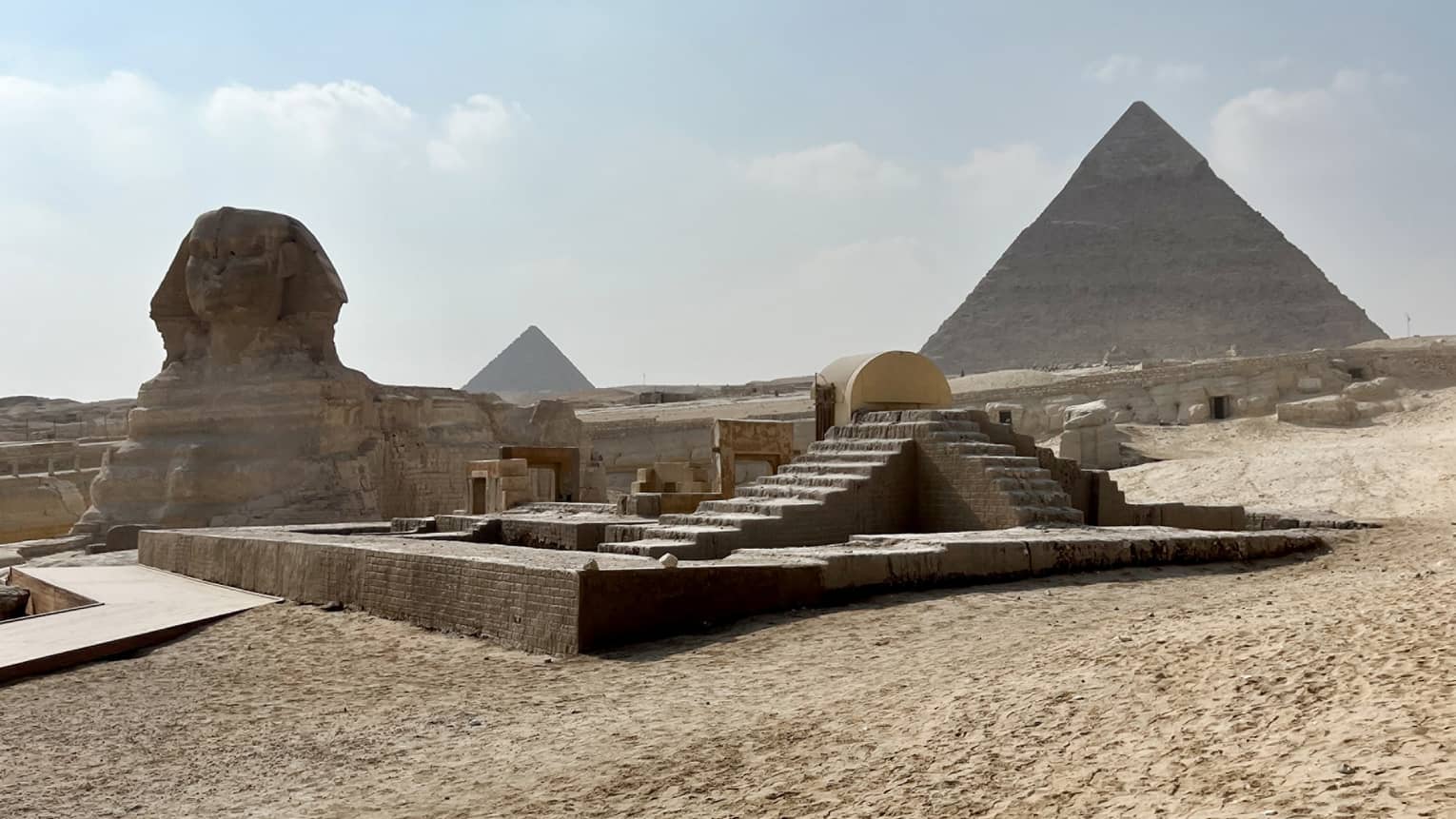 Distant view of the sphynx, desert sand and stone steps leading up to two pyramids under blue sky with fluffy clouds.