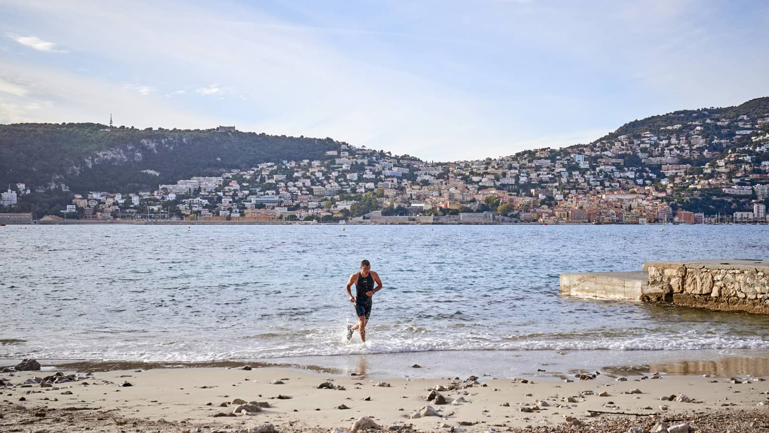 Man jogging in water toward sandy shore, hills in backdrop