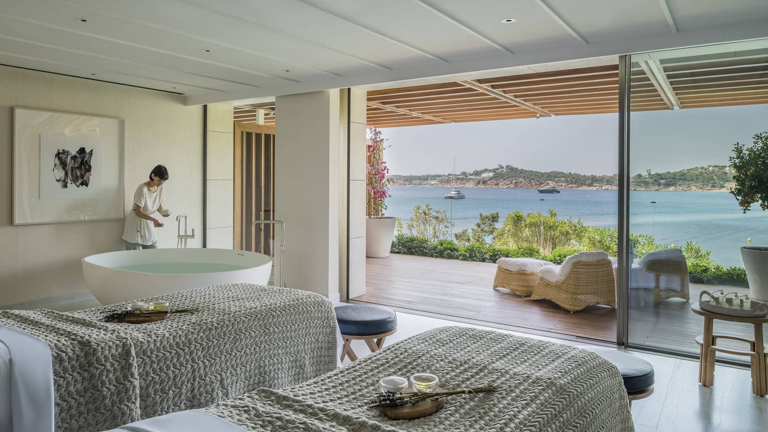 A four seasons staff works in a room with two side by side spa tables draped with gray linens face an open veranda overlooking a body of water in Greece