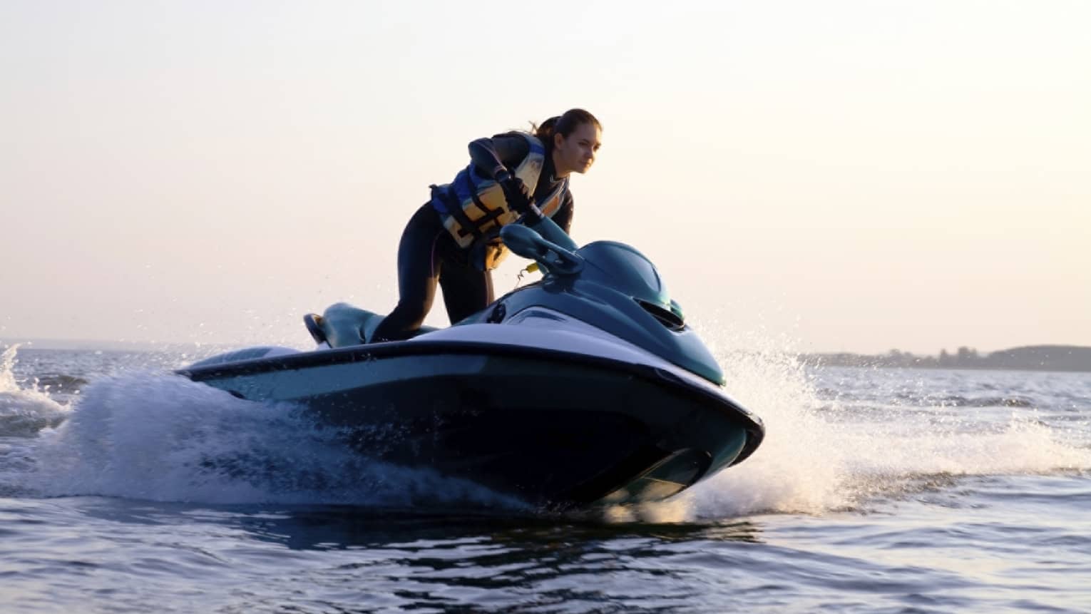 Woman wearing life jacket stands, steers jet ski on ocean at sunset