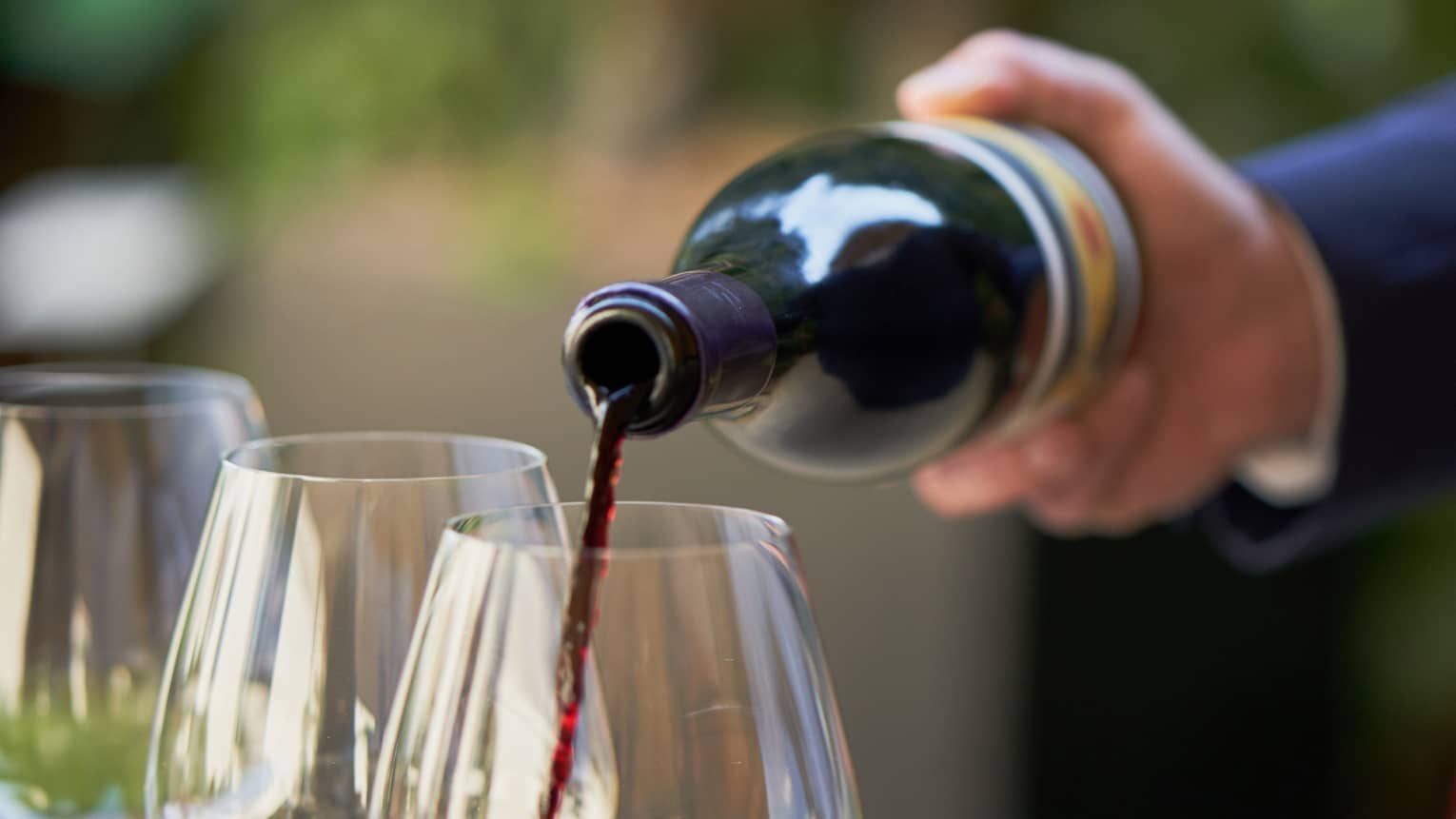 A close up of hands pouring a bottle of red wine into a clear wine glass that rests on a wooden tray, outdoors