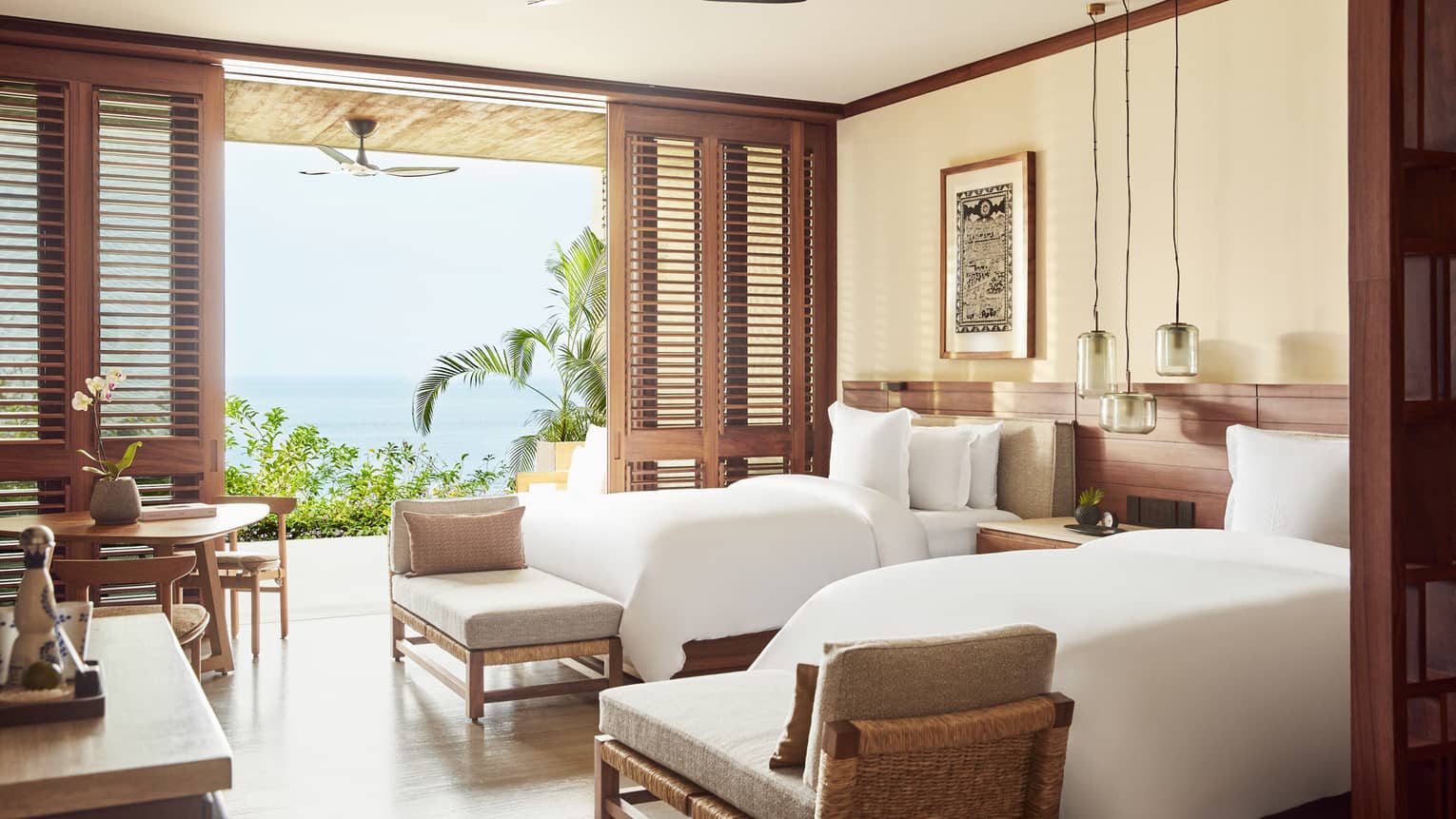 A beachfront room with 2 beds, a small table, benches at the end of each bed and a large open door to outside.