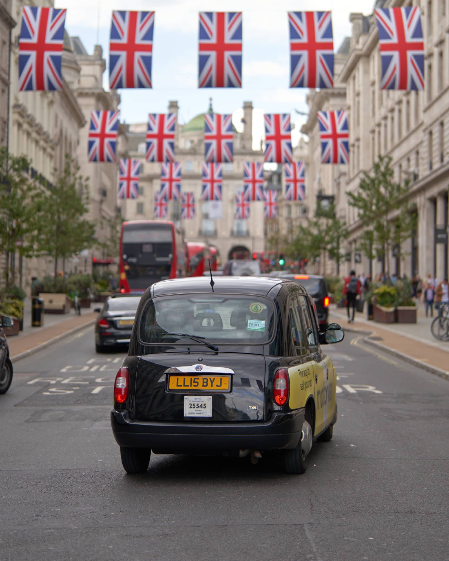 Rear view of London-style black cab, double-decker buses and other cars on road strung across with rows of Union Jack flags.