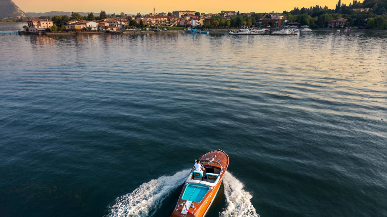 Person driving a wooden speedboat across a lake towards shore at sunset