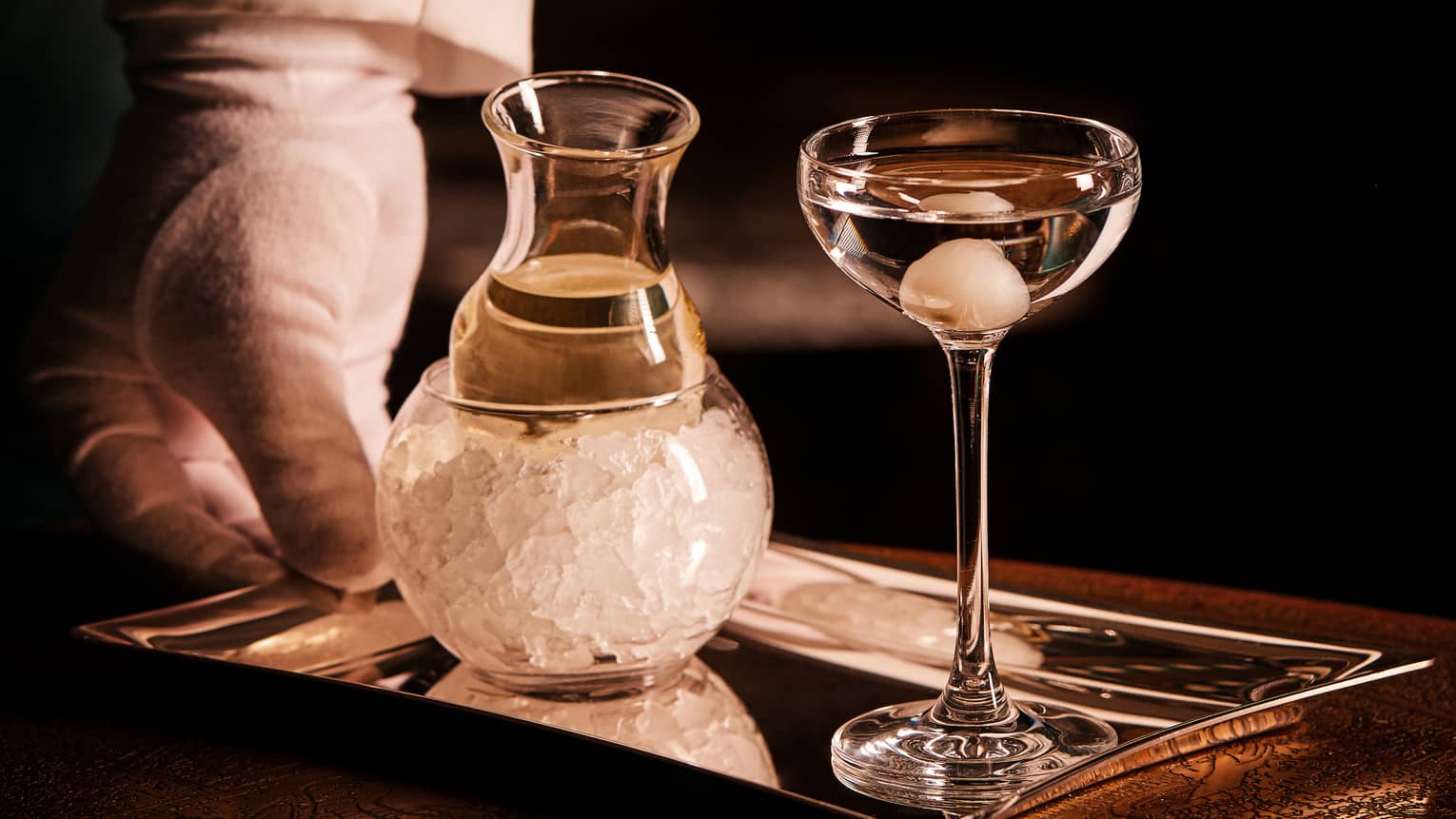 Gloved hand holds a small, mirrored tray with a dainty ice-chilled carafe and a full stemmed, onion-garnished cocktail glass.