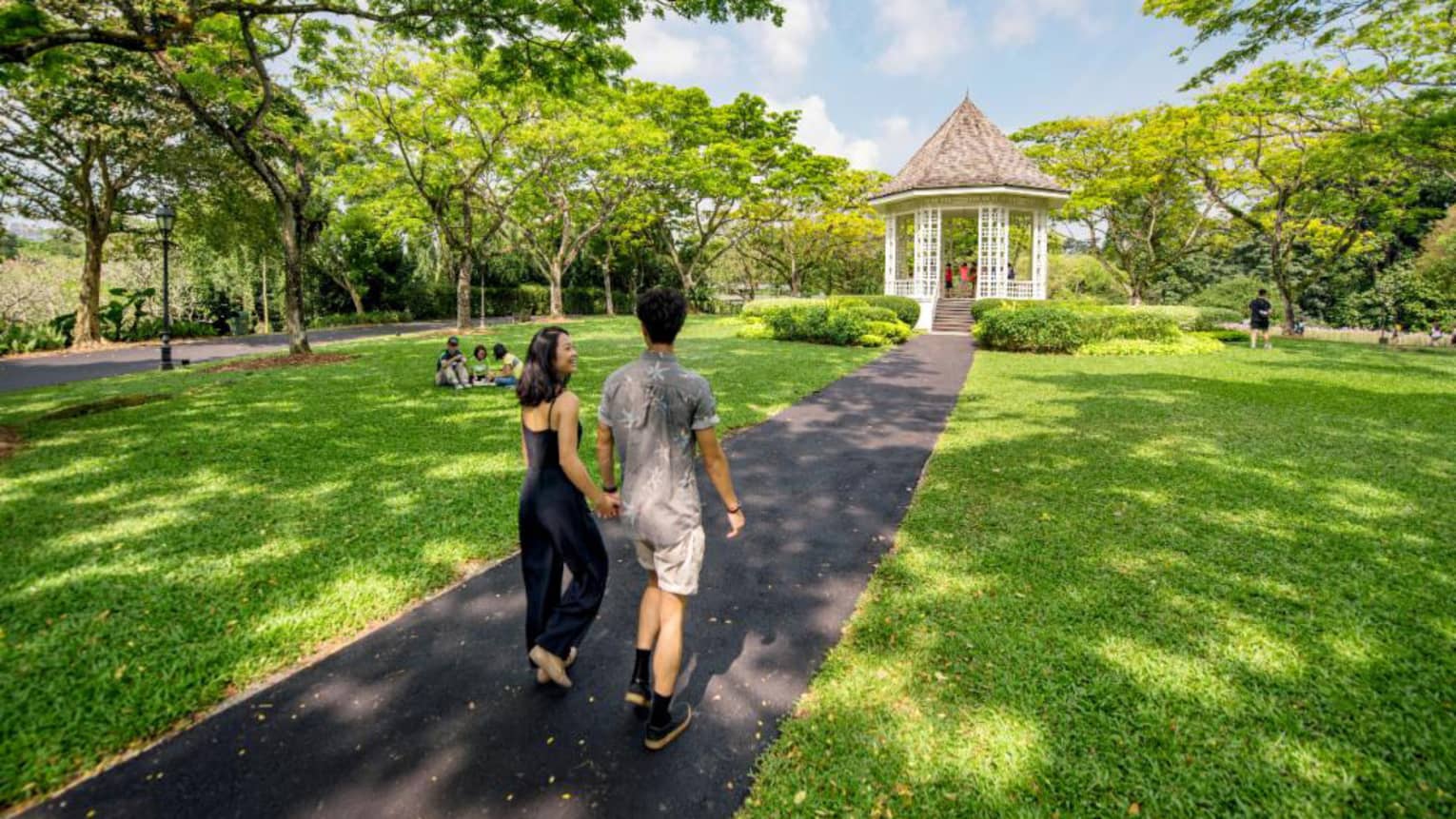A man and woman hold hands and walk along an asphalt trail through the Singapore Botanic Gardens 