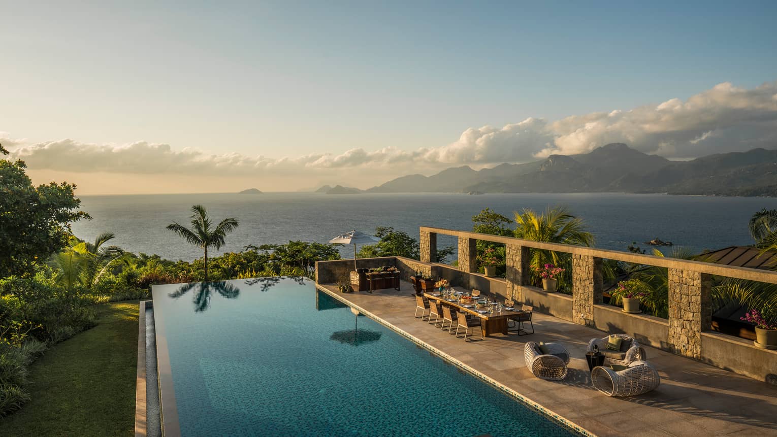 Infinity pool overlooking ocean with palm trees, table, chairs and lounge area 