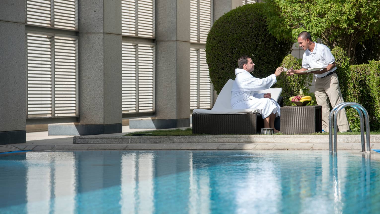 Man in white bathrobe sits on lounge chair on sunny swimming pool deck by fruit bowl, hotel stands hands him cloth