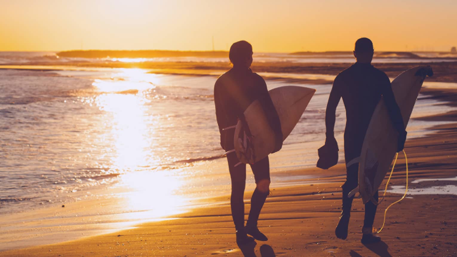 Silhouettes of two people carrying surfboards across beach at sunset