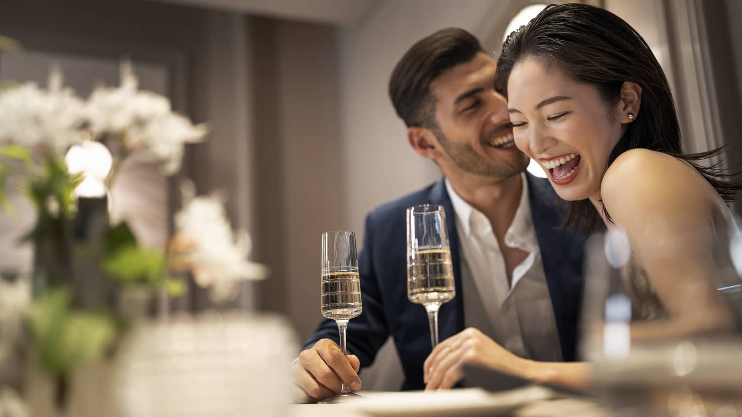 A couple sharing a meal on a table adorned with white flowers, holding champagne glasses and engaged in conversation and laughter.