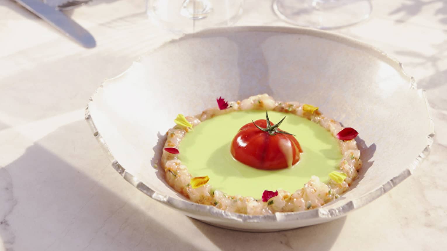 Green gazpacho surrounded by shrimp with halved tomato in centre in large ecru bowl on dining table