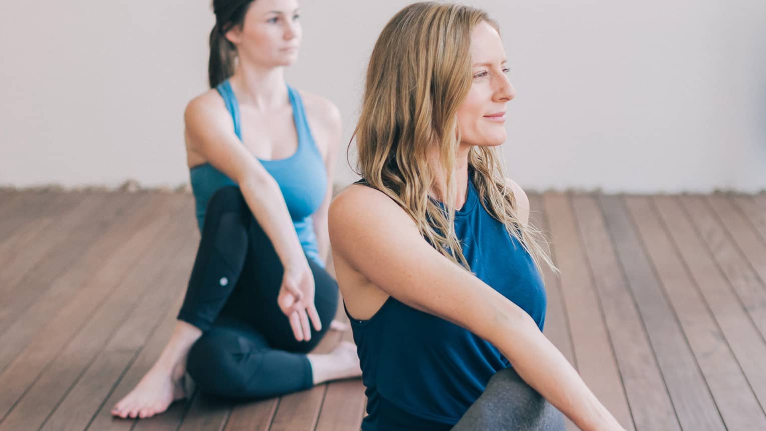 One blonde woman and one brunette woman sit with one leg crossed over the other with their bodies turned to one side and their front hands forming a lam mudra