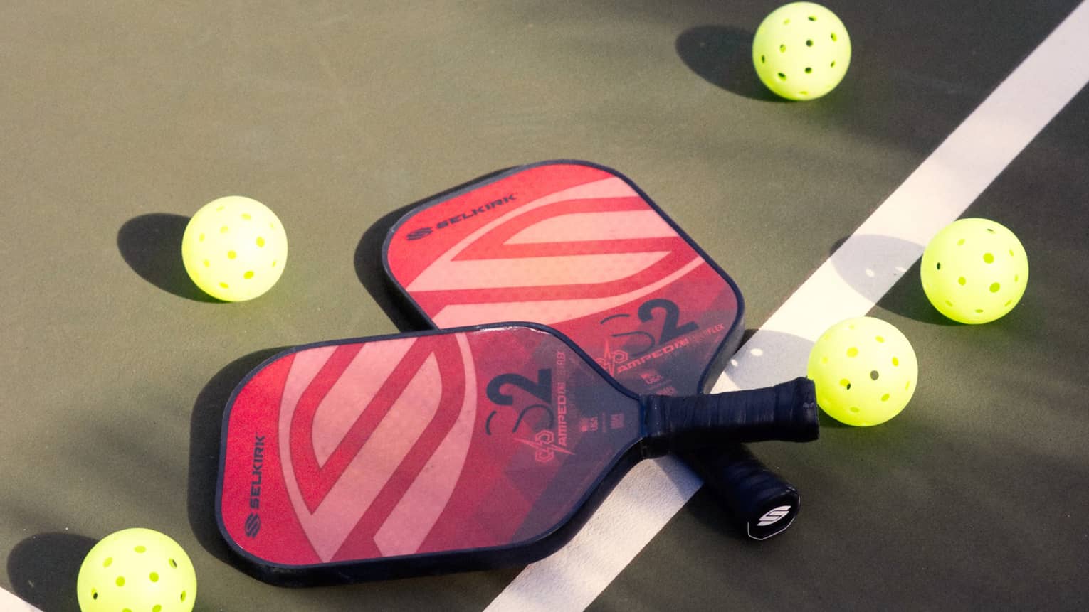 Close-up of two red and pink pickleball racquets, handles crisscrossed on a white court line, surrounded by five pickleballs.