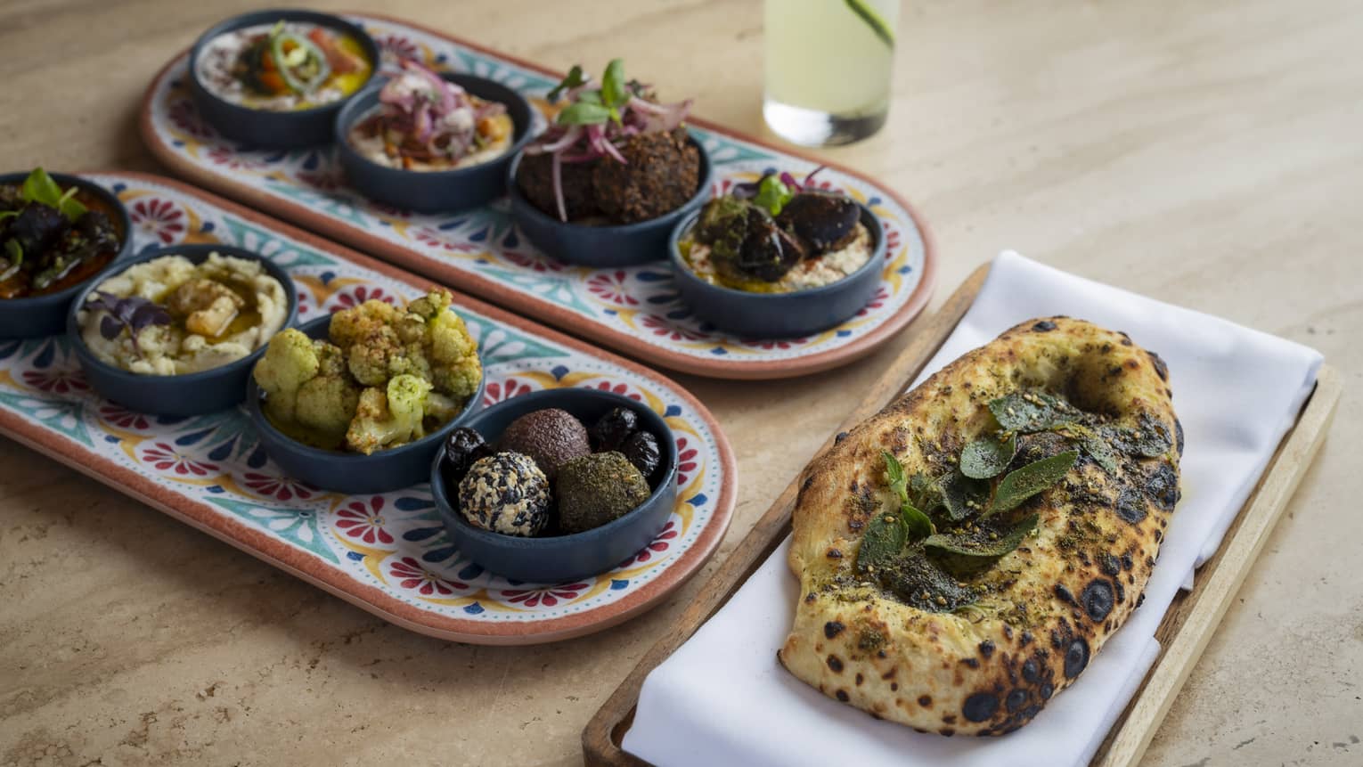 Two platters lined with four small plates of Mediterranean dishes and a platter of fresh-baked bread topped with leaves on a light-wood table