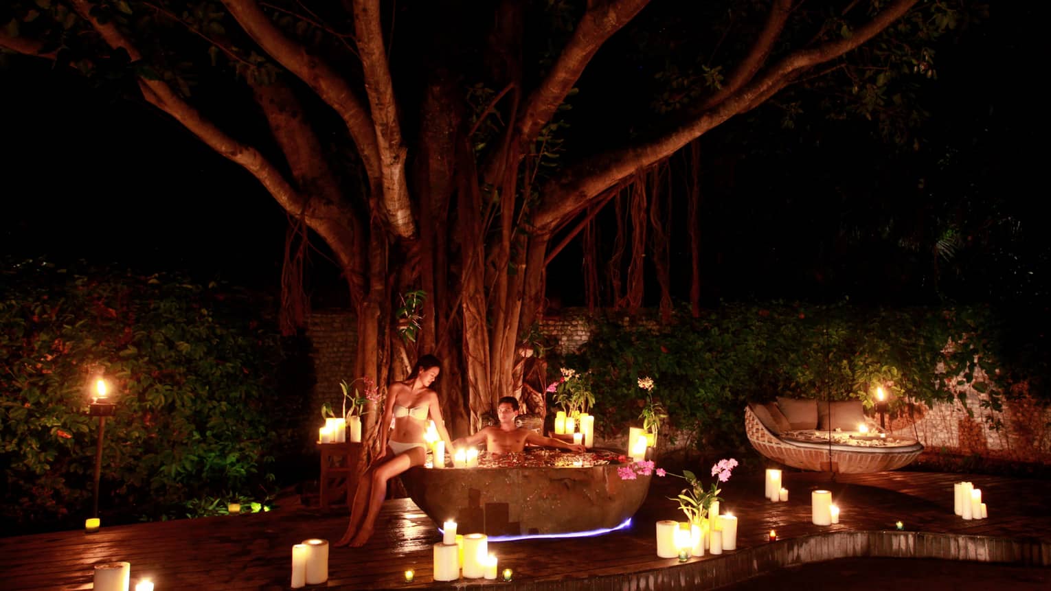 Man in outdoor whirlpool tub at night, woman in bikini on edge, surrounded by lit candles