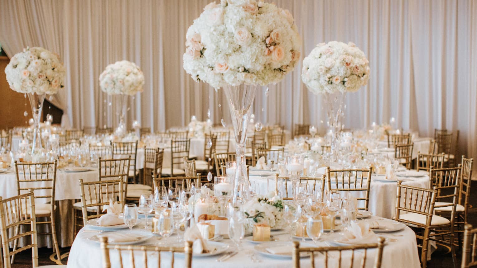 Round tables with white table cloths, wood chairs, white flowers, and white table settings.