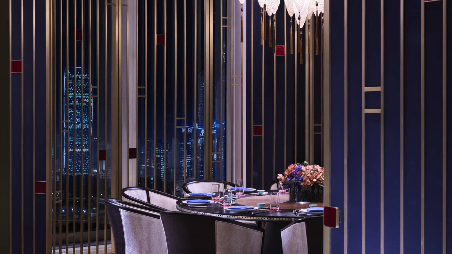 Elegant round dining table, chairs under small crystal light in front of window with city views at night