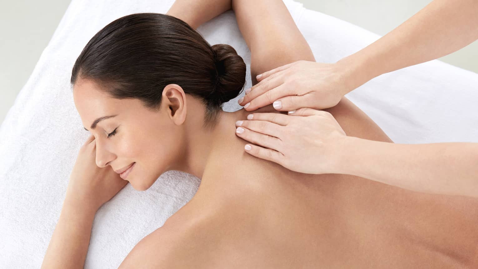 Woman smiles, lies on massage table as hands massage bare shoulders