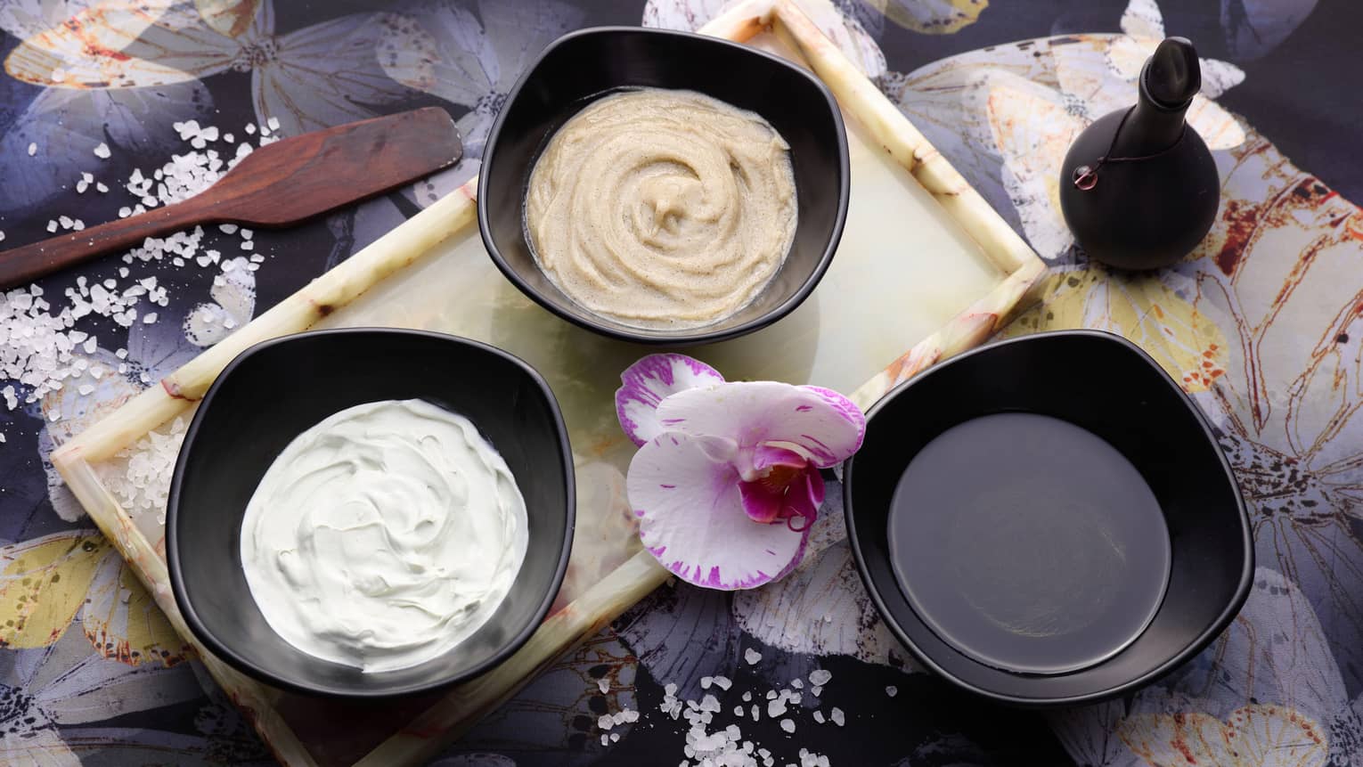 Looking down at three small black bowls with lotions, oils, fresh orchid flower