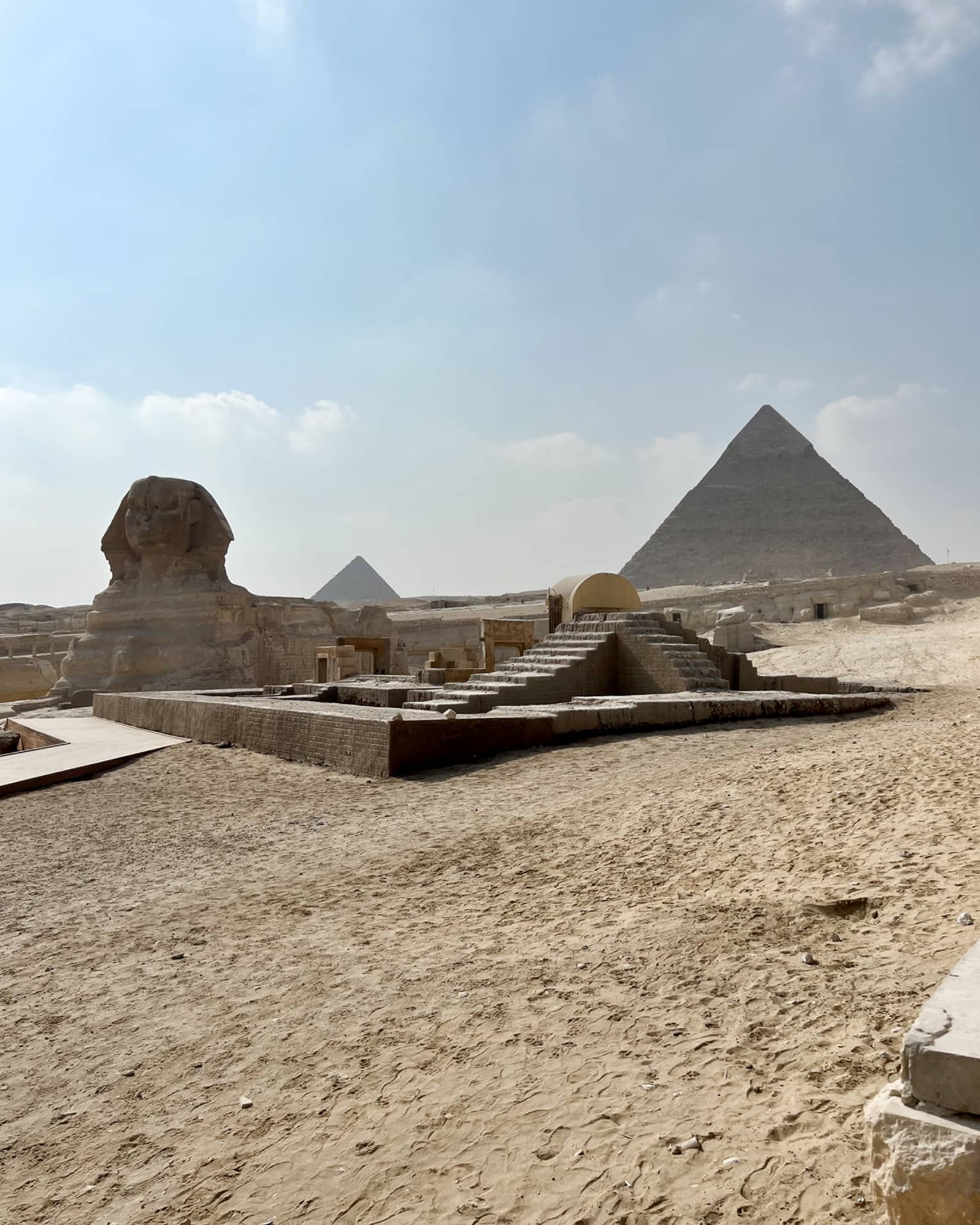 Distant view of the sphynx, desert sand and stone steps leading up to two pyramids under blue sky with fluffy clouds.