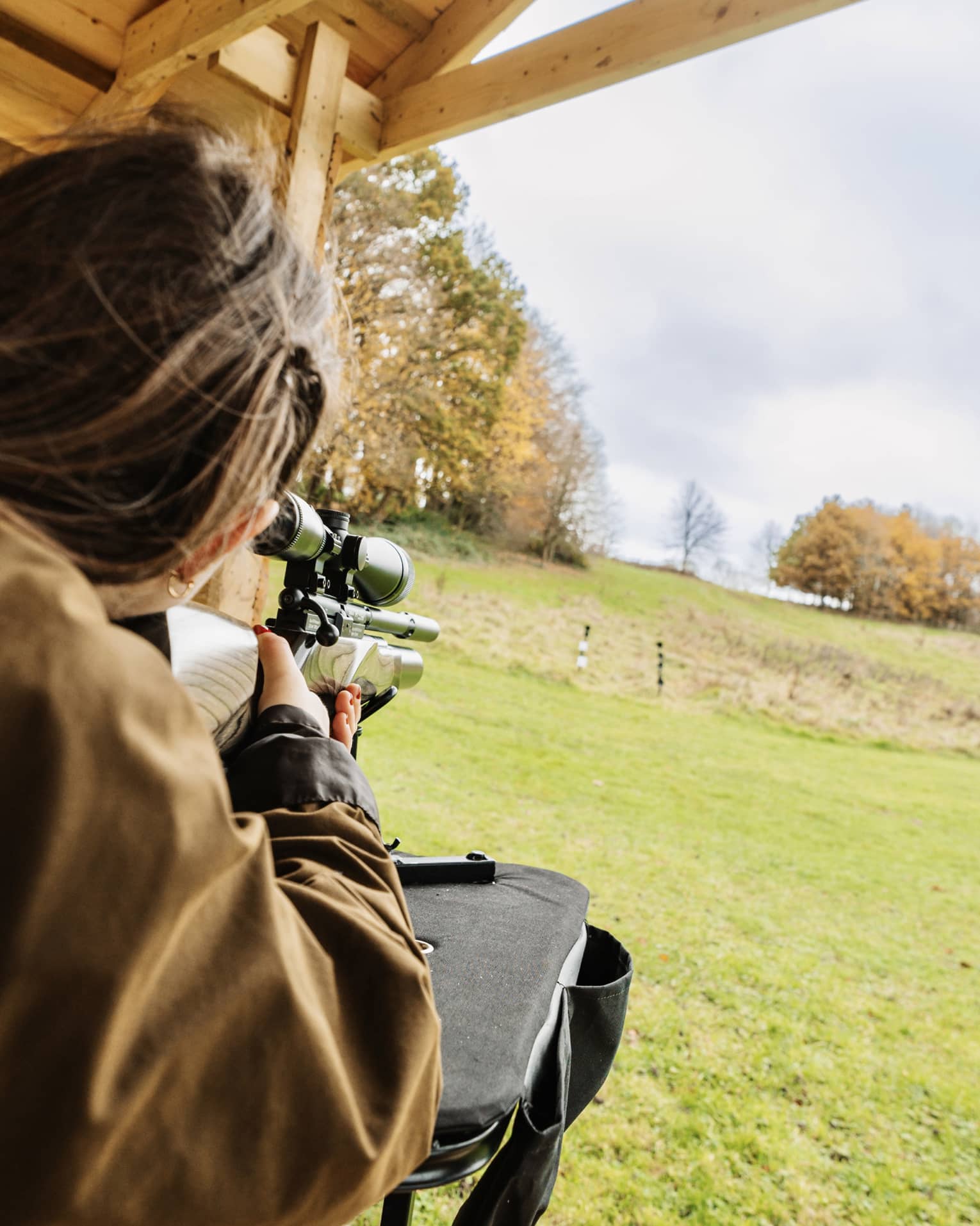Rear view of a person in a brown jacket aiming an air rifle at small, distant targets in a large field amid autumn trees.
