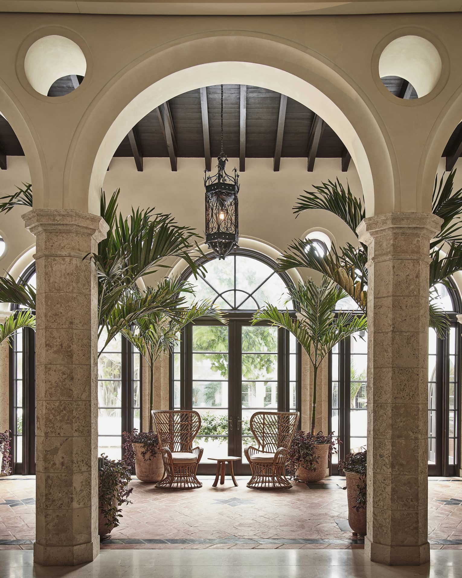 An archway looking at two wood chairs by a large window and small table, there are palm trees around the chairs.