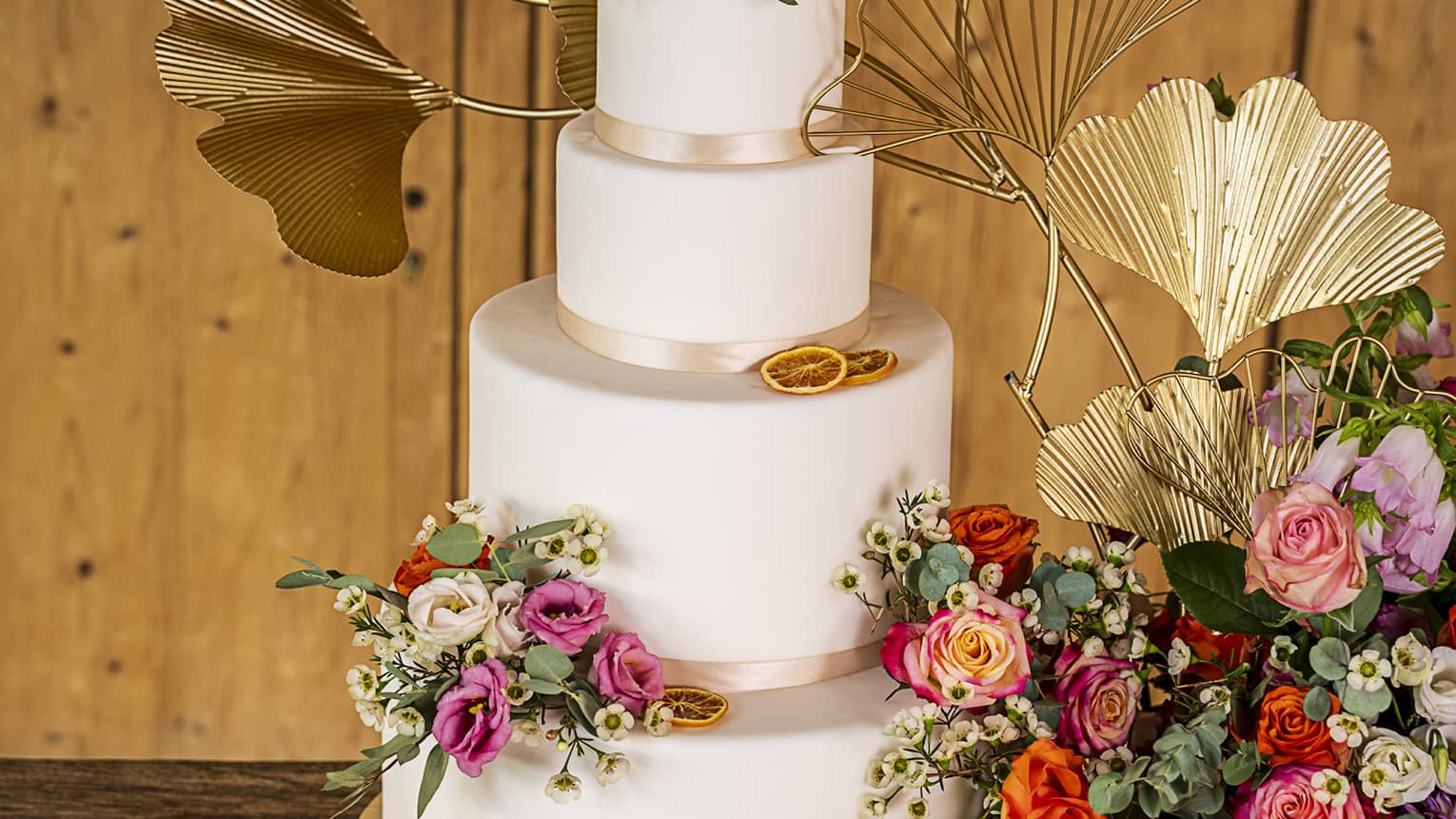 Three-tiered wedding cake with autumn-coloured flower and gold metal gingko accents beside large bouquet